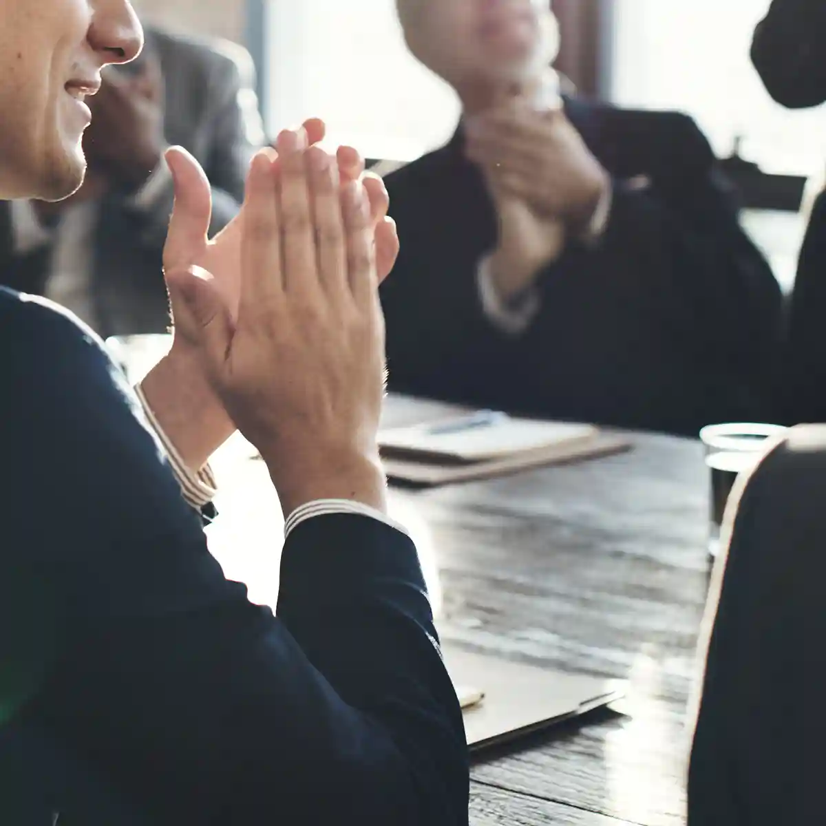 workshop-fuehrungskraefte-austausch-wit-desktop Ein Mann in einem Anzug applaudiert während einer Besprechung mit anderen Anwesenden im Hintergrund.